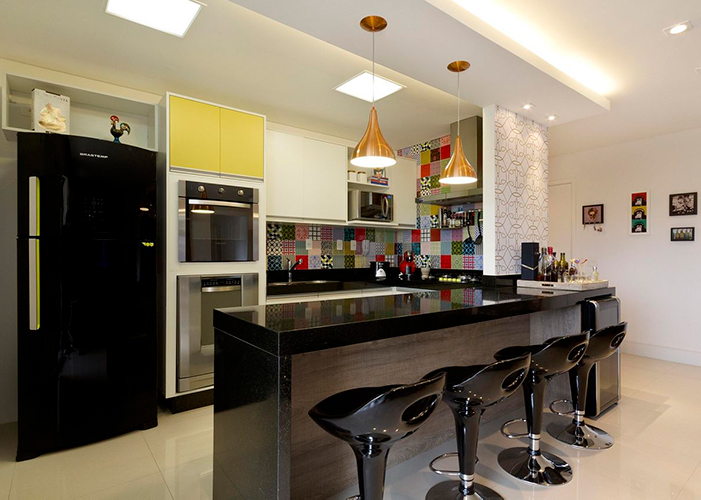 cozinha americana, com detalhe na parede com azulejos quadradinhos coloridos, geladeira preta, bancada e banquetas giratórias pretas com encosto.