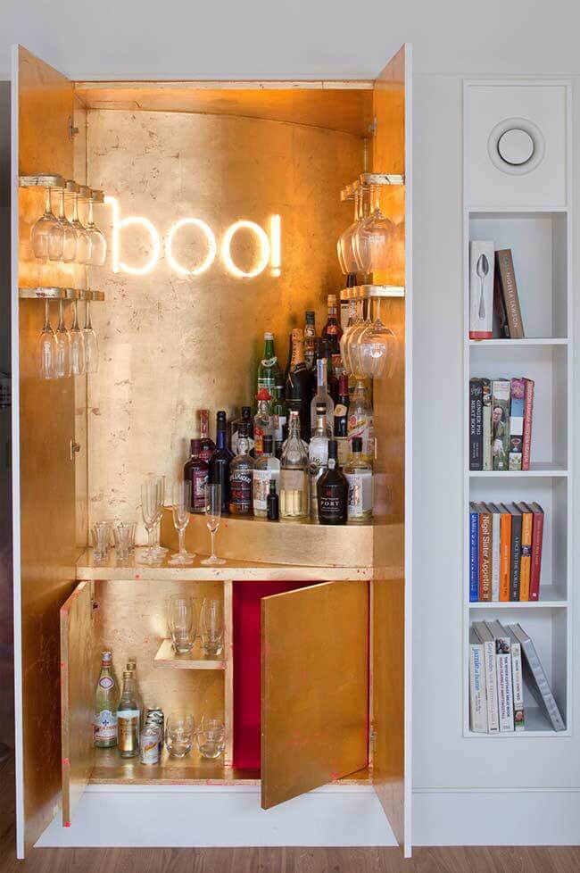 Bar embutido na parede, com as portas abertas mostrando o conteúdo que é taças, copos e bebidas. Ao lado nichos com livros 