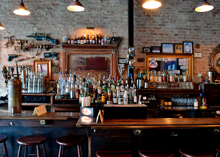  Estrutura interna de um bar com balcão e banquetas de madeira, muitas bebidas. Parede de tijolinhos e pendentes
