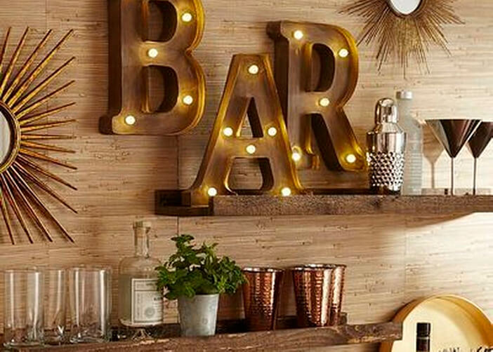 Luminária pendurada na parede em formato de letra formando a palavra do bar. Prateleiras apoiando copos e adornos