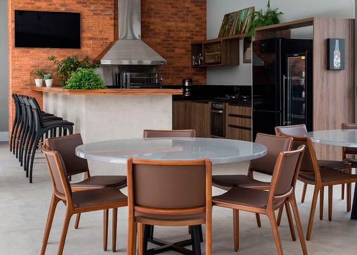 Espaço gourmet em casa com 2 mesas redondas, cadeiras de madeiras estofadas, ao fundo pia em madeira, balcão com baquetas e eletrodoméstico preto