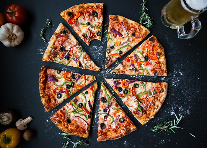 Pizza de queijo, tomate, pimentão verde, azeitona roxa fatiada em um fundo preto ao redor decorando: alho, pimentão, alecrim e um copo com chopp