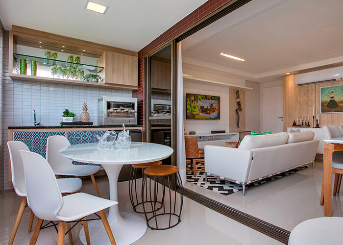 varanda integrada com sala e cozinha. Mesa branca redonda com cadeiras brancas, churrasqueira elétrica em cima da bancada