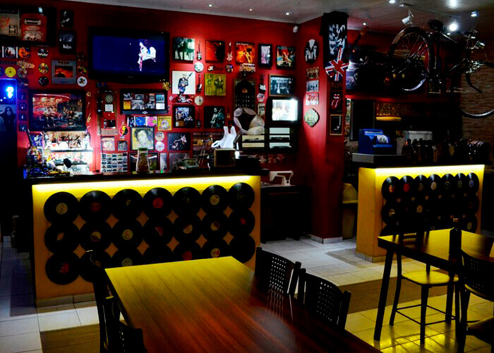 Ambiente de bar com mesa de madeira e cadeiras pretas, paredes vermelha com vários quadros e objetos vintages pendurados