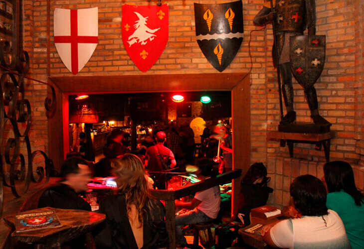Bar com pessoas sentadas em mesas feitas de troncos de madeira e ferro. Paredes de tijolos com quadros em formato de escudos e uma armadura de cavaleiro no canto direito superior