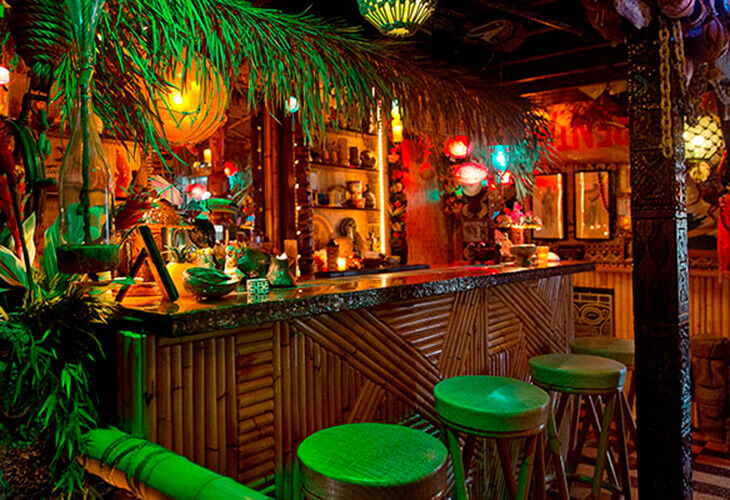 Balcão de bar revestido de bamboo, com banquetas altas. Muitas folhagens e uma coluna de apoio esculpida em formato de toten