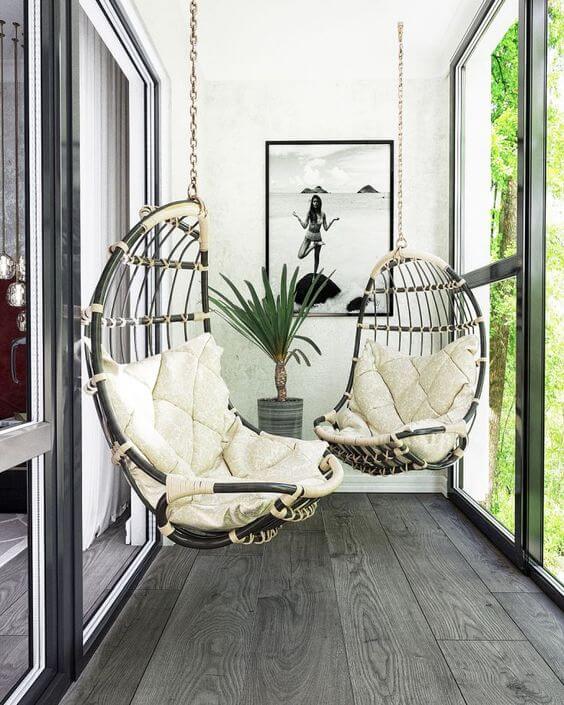 varanda com piso de madeira cinza, e duas cadeiras suspensas na cor bege, quadro na parede