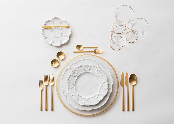 mesa posta vista de cima com pratos detalhados, talheres dourados e taças.