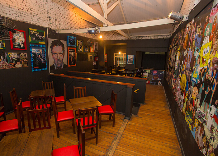 Bar interno com parede forrada de personagens de seriados, mesas de madeira quadradas com cadeiras