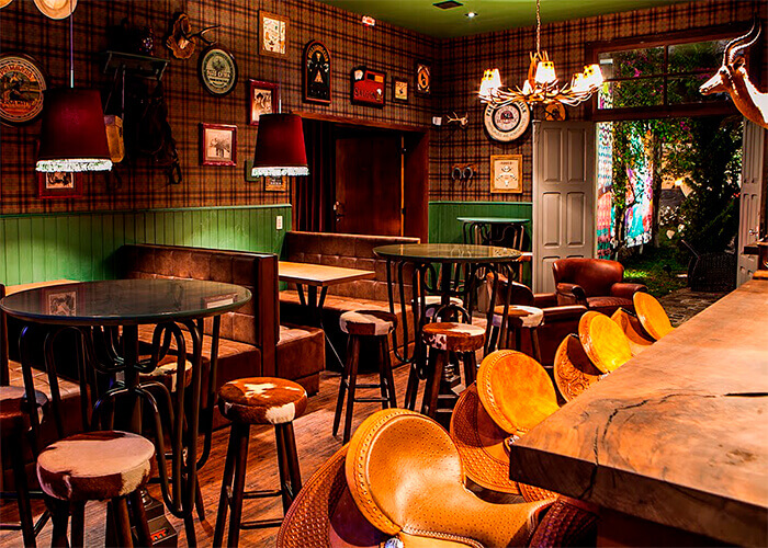 Bar decorado com balcão rústico de madeira, banquetas de sela de cavalo, parede metade de madeira verde, e outra com papel de parede xadrez com vários quadros