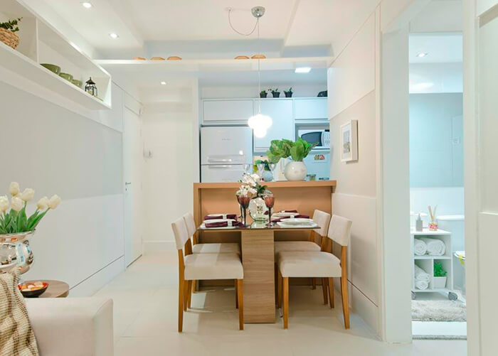 mesa central de jantar com 4 cadeiras com pés em madeira, ao fundo geladeira branca e iluminação branca