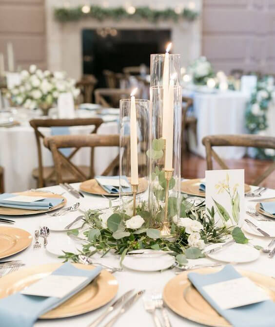 mesa redonda com toalha clara, pratos amarelos e guardanapo azul claro, com arranjo central com velas