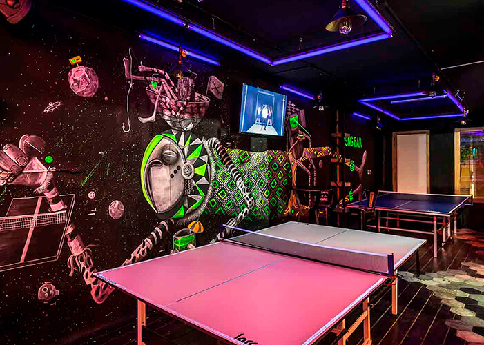 ambiente com paredes pretas. 2 mesas de ping pong com desenhos neons na parede