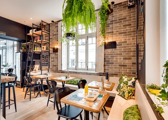 ambiente interno da cafeteria com mesas personalizadas, parede de tijolinho e booth com almofadas