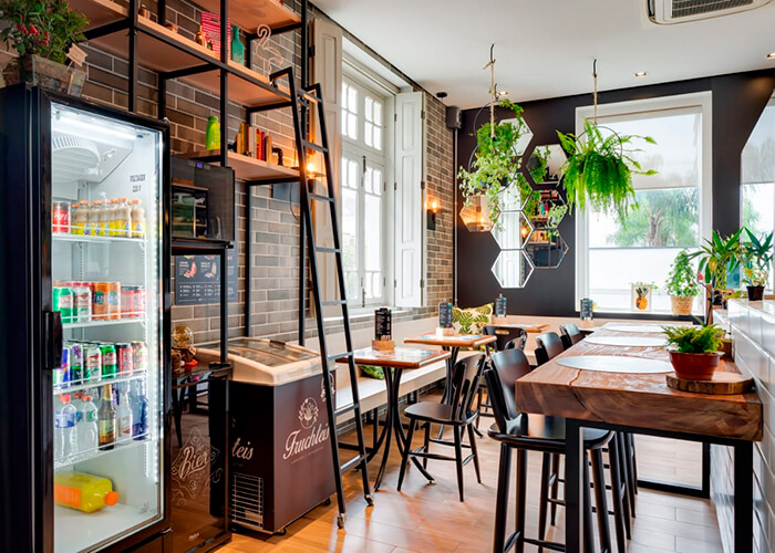 ambiente interno de cafetería com plantas para decoração, parede de tijolinhos, mesas de madeira.