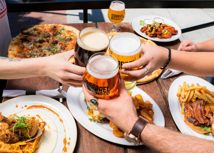 mãos-de-homem-brindando-com-cerveja-sobre-mesa-com-diversas-comidas