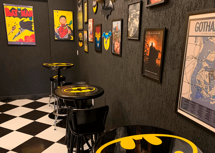 Ambiente com parede preta com quadros decorativos e chão quadriculado, mesas personalizadas com o símbolo do Batmam.