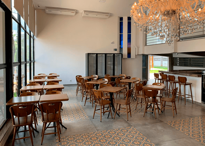 Cafeteria com vários conjuntos de mesas e cadeiras em madeira. Um grande lustre se destaca.