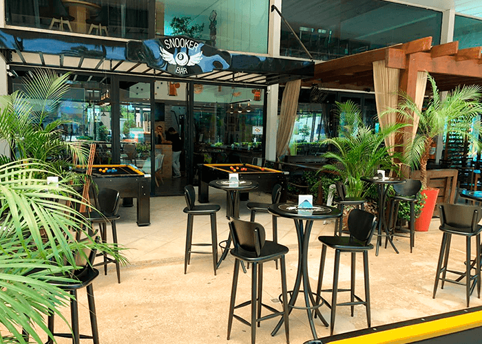 ambiente externo com mesas altas e banquetas. Várias plantas